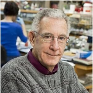 Thomas N. Seyfried shoulder and head image appears sat in laboratory wearing a grey jumper and purple shirt with someone working in the background