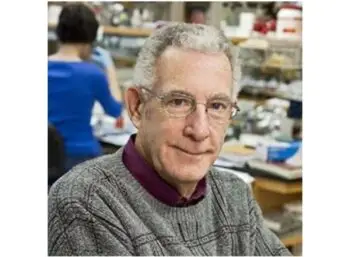 Thomas N. Seyfried shoulder and head image appears sat in laboratory wearing a grey jumper and purple shirt with someone working in the background